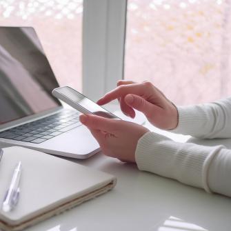 Tekoälyllä teetetty kuvituskuva, jossa henkilö kannettavan tietokoneen ja puhelimen äärellä.