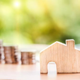 Kuvituskuva, jossa puinen talokoriste ja taustalla kolikkopinoja.