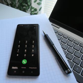 Puhelin, tietokone ja muistilehtiö pöydällä.