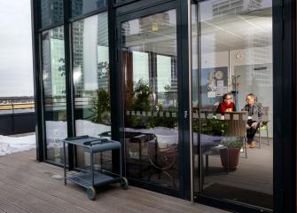 Lasiseinäinen huone, jossa kaksi iäkästä henkilöä istuu pöydän ääressä keskustelemassa; ulkopuolella näkyy terassi, lumisia kohtia ja kaupunkimaisema taustalla.