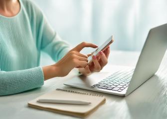 Henkilö käyttää kännykkäänsä kannettavan tietokoneen äärellä.