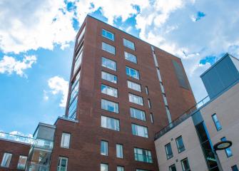 Punatiilinen kerrostalo kirkkaassa päivänvalossa, taustalla sininen taivas ja valkoisia pilviä. Rakennuksessa on useita ikkunoita ja sen vieressä näkyy osia muista moderneista rakennuksista.