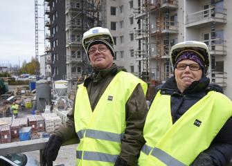 Kaksi henkilöä rakennustyömaalla, yllään keltaiset turvaliivit ja suojakypärät, taustalla kerrostalojen rakentaminen ja rakennusmateriaaleja.